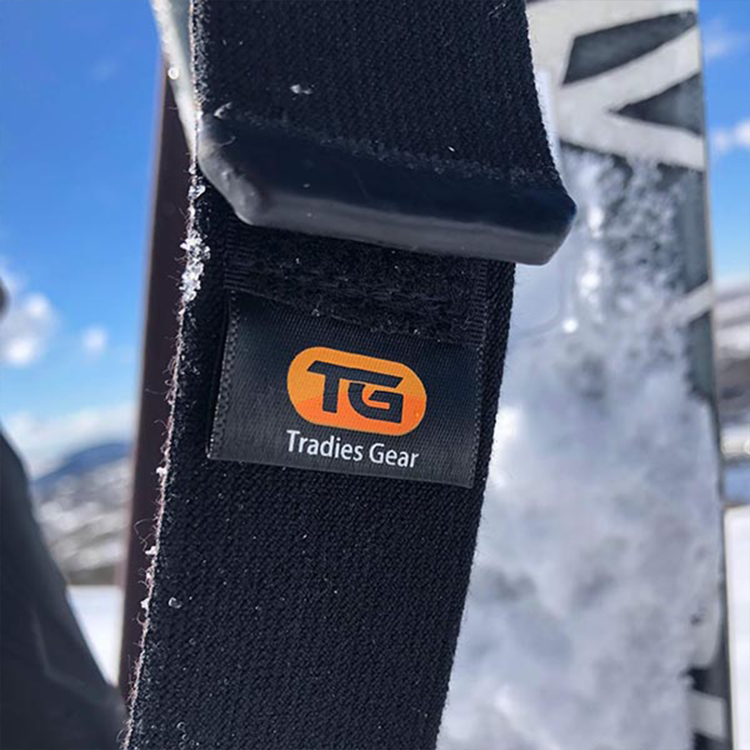 Close-up of Tradies Gear no-buckle belt showing brand tag TG with snow-covered background. snowboarding and skiing belt